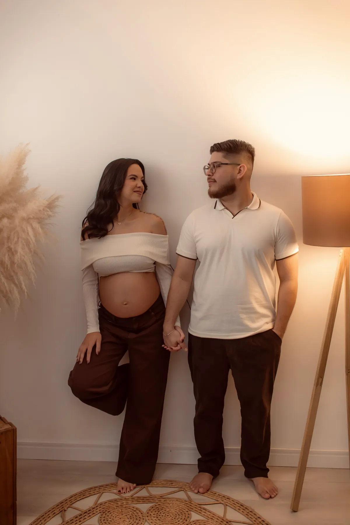 Casal de mãos dadas no ensaio gestante, ao lado de luminária e capim-dos-pampas