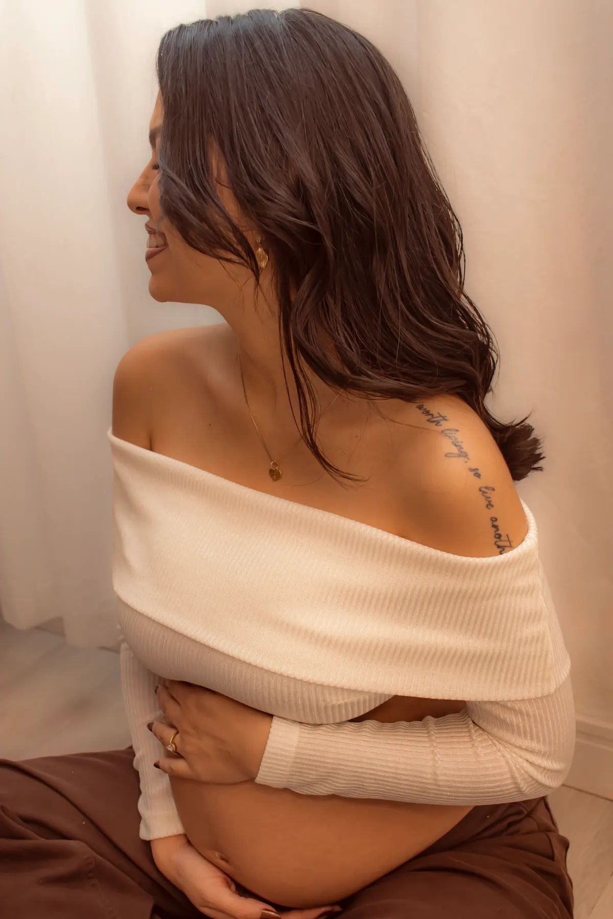 Gestante de perfil com blusa ombro a ombro e as mãos sobre a barriga
