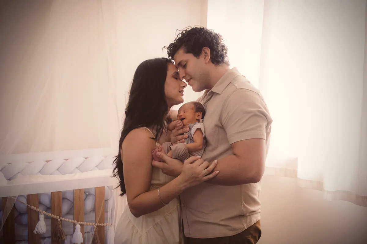 Casal com o bebê recém-nascido no colo, contra a luz suave da janela